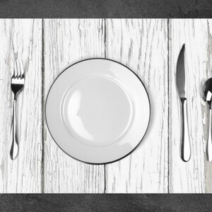 Puede incluir: Un plato de cena de cer&aacute;mica blanca con un borde negro se encuentra sobre una mesa de madera blanqueada. Un tenedor, un cuchillo y una cuchara se colocan alrededor del plato.