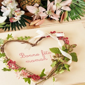 Peut inclure: Une couronne en forme de cœur faite de vignes et de verdure avec des baies rouges et un ruban blanc. La couronne est sur une carte beige avec le texte "Bonne fête maman!" en rose.