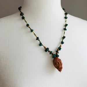 May include: A necklace featuring a carved wooden pendant with a detailed face design. The necklace has a beaded chain with green and brown accents, displayed on a white mannequin.
