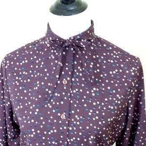 May include: A long-sleeved button-down shirt with a brown background and a white, blue, and green polka dot pattern. The shirt has a high collar and a tie at the neck.