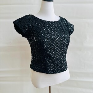 May include: A black sequined top with short sleeves. The top is displayed on a white mannequin. The sequins create a textured, chevron-like pattern across the fabric. The neckline is a simple scoop. The top is a classic, elegant style.
