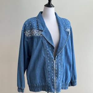 May include: A light blue denim jacket with a silver zipper closure. The jacket features decorative silver and white embellishments on the shoulders and down the front. The jacket has long sleeves and a banded waist and cuffs.