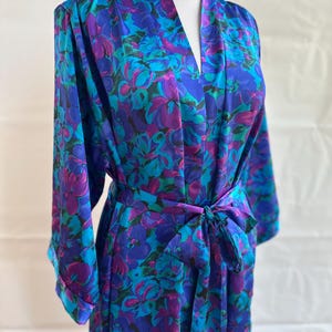May include: A floral-patterned robe in shades of blue, teal, and purple, tied at the waist with a matching sash. The robe features a V-neck and long sleeves with turned-up cuffs. The fabric has a silky sheen.