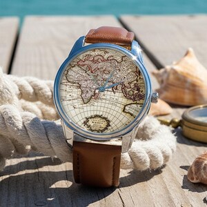 Pode incluir: Um relógio de pulso com um design de mapa-múndi no mostrador e uma pulseira de couro castanho. O relógio está exposto numa superfície de madeira com conchas, uma bússola e uma corda, com o oceano azul ao fundo.