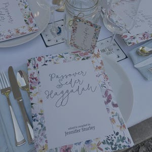 Op de afbeelding: Een wit boekje met bloemenpatroon voor de Pesach Seder Haggadah met de titel "Passover Seder Haggadah" en de tekst "bewerkt en samengesteld door Jennifer Sturley".