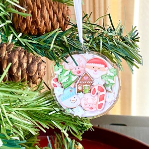 May include: A clear, round Christmas ornament with a festive design. The ornament features gingerbread cookies shaped like Santa, a snowman, reindeer, and a gingerbread house, with red, green, and white accents. It hangs from a white ribbon on a Christmas tree.
