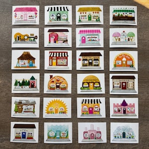 May include: A collection of 24 square watercolor illustrations of various storefronts, including a cafe, bookstore, and tattoo parlor. Each miniature building features unique architectural details and colorful awnings, set against a neutral background.