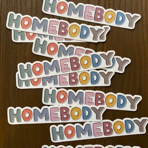 May include: A stack of stickers with the word "Homebody" written in a variety of colors, including pink, blue, yellow, green, and brown. The stickers are arranged in a staggered pattern, with the top sticker being the largest and the bottom sticker being the smallest.