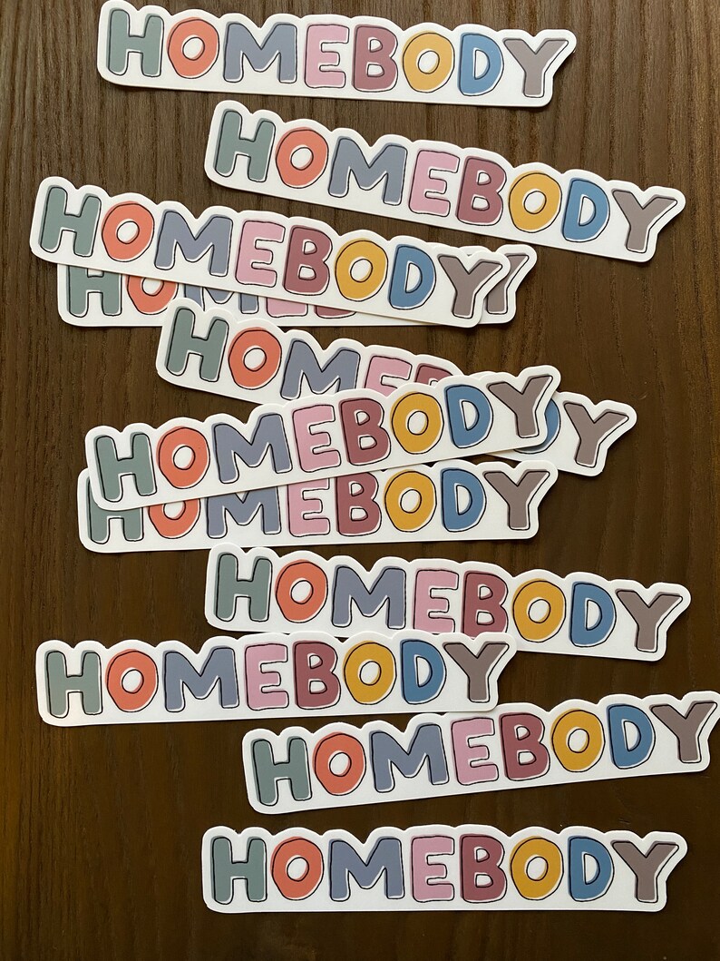 May include: A stack of stickers with the word "Homebody" written in a variety of colors, including pink, blue, yellow, green, and brown. The stickers are arranged in a staggered pattern, with the top sticker being the largest and the bottom sticker being the smallest.