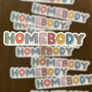 May include: Multiple stickers with the word "HOMEBODY" in colorful, outlined letters. The stickers are stacked on top of each other, with the top sticker being the most in focus.
