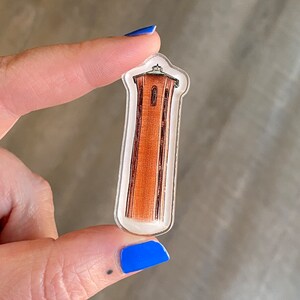 May include: A clear acrylic charm featuring a detailed illustration of a red brick tower with a pointed roof. The tower has a small window and is set against a white background.
