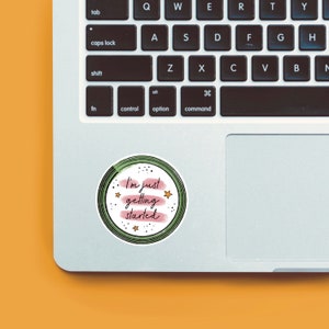 May include: A round sticker with a green and white border and a pink background. The sticker has white stars and the text "I'm just getting started" in black.