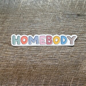 May include: A sticker with the word "HOMEBODY" in colorful letters. The letters are outlined in white and have a slightly raised, 3D effect.