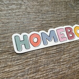 May include: A sticker with the word "HOMEBODY" in colorful, outlined letters. The letters are in different colors, including green, orange, blue, pink, and yellow.