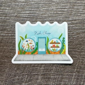 May include: A sticker featuring a whimsical illustration of a fish shop. The design includes a light blue facade with a door, two circular windows displaying fish and aquatic plants, and the words "Fish Shop" in cursive.
