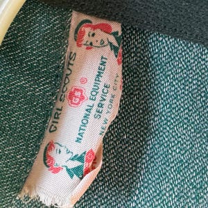 May include: A white fabric tag with green trim and red accents. The tag reads "Girl Scouts" and "National Equipment Service, New York City". The tag features a cartoon image of a girl scout.