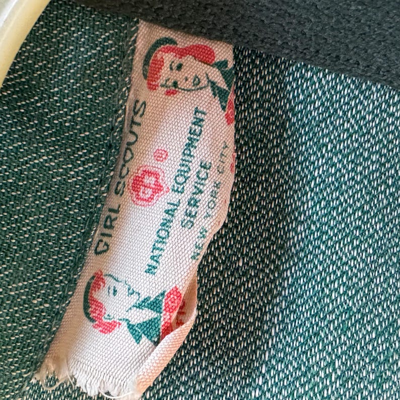 May include: A white fabric tag with green trim and red accents. The tag reads "Girl Scouts" and "National Equipment Service, New York City". The tag features a cartoon image of a girl scout.