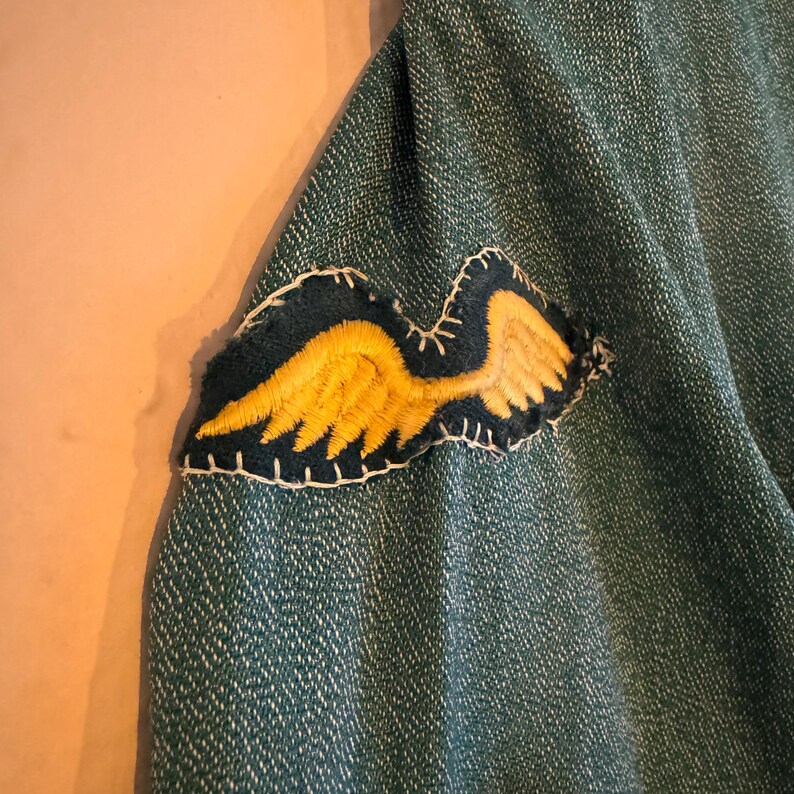 May include: A green denim jacket with a yellow embroidered wing patch sewn onto the sleeve.