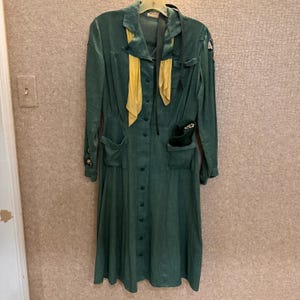 May include: A vintage green Girl Scout uniform dress with a yellow sash and a black belt. The dress has a button-up front and two pockets on the front. The dress has a patch on the left sleeve that says "4".