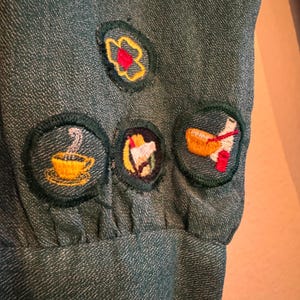 May include: Green fabric with four embroidered patches. The patches are circular and feature a flower, a cup of coffee, a bird, and a spoon and bowl.