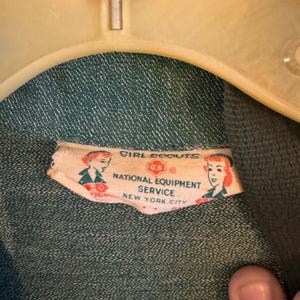 May include: A close-up of a green fabric label with white text that reads "Girl Scouts National Equipment Service New York City". The label has a red and white logo with a circle and a cross inside. There are two small illustrations of girls in scout uniforms on the label.