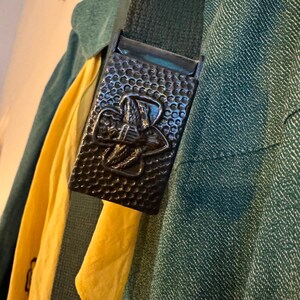 May include: A dark green fabric belt with a metal buckle featuring a Girl Scout logo. The buckle is textured and has a raised design.