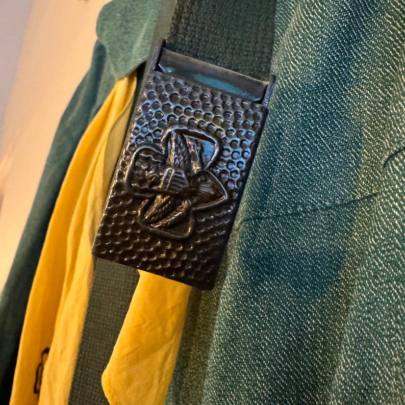 May include: A dark green fabric belt with a metal buckle featuring a Girl Scout logo. The buckle is textured and has a raised design.