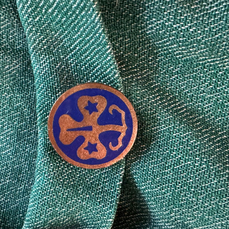 May include: A blue and gold Girl Scout pin with a four-leaf clover design and three stars.