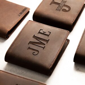 May include: Five brown leather wallets with different personalized engravings. The wallets are arranged in a staggered pattern on a white surface. The engravings include initials, names, and a deer head design.
