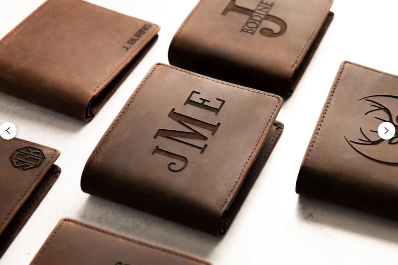 May include: Five brown leather wallets with different personalized engravings. The wallets are arranged in a staggered pattern on a white surface. The engravings include initials, names, and a deer head design.