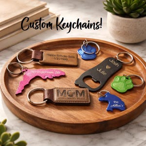 May include: A collection of custom keychains in various shapes and colors, including a pink Florida outline, a paw print, and a bottle opener. Some keychains have personalized text, such as names and dates. The text "Custom Keychains!" is displayed.