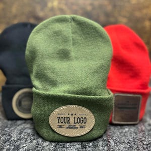 May include: Three beanies in black, green, and red. The green beanie is in focus, displaying a leather patch with the text "YOUR LOGO" and "LASTING IMPRESSIONS 2014". The other beanies have similar patches. The beanies are on a speckled surface.