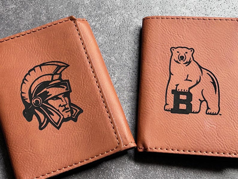 May include: Two brown leather wallets with different logos. The first wallet has a black and white logo of a Roman soldier wearing a helmet. The second wallet has a black and white logo of a bear holding a letter "B".