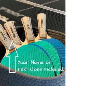 May include: A basket holds four ping pong paddles with wooden handles labeled "Chopper", "Smasher", "Ace", and "Spinner". The paddle faces are teal and blue. The text "Your Name or Text Goes included" is also visible.
