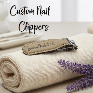 Puede incluir: Un cortaúñas personalizado con un mango de cuero color beige, grabado con el texto "Jessica Nailed It!". Descansa sobre una toalla blanca enrollada. La imagen incluye una ramita de lavanda morada y el texto "Custom Nail Clippers".