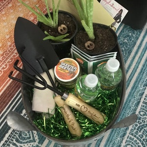 May include: A galvanized metal bucket filled with green and white shredded paper, two clear plastic water bottles, a small jar of insect repellent labeled "Skeeter Beeter", two potted aloe vera plants, a small trowel, a hand rake, and a wooden gardening tool with the text "Samantha & Nick" and "Happy Gardening!" engraved on the handle.