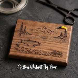 May include: A rectangular walnut wood fly box featuring a laser-engraved scene of a mountain landscape, a lake, a fish, and a crayfish. A yellow and black fishing fly rests on the surface. The text Custom Walnut Fly Box is printed below.