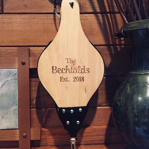 May include: A wooden bellows with a black leather handle and a silver tip. The bellows is engraved with "The Bechtolds Est. 2018".