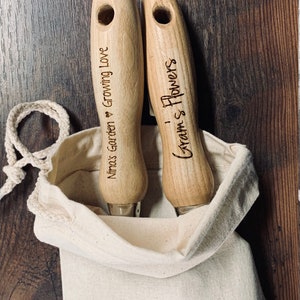 May include: Two wooden gardening tools with handles engraved with "Nimos Garden Growing Love" and "Gram's Flowers" in a white canvas bag.