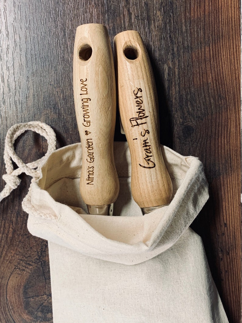 May include: Two wooden gardening tools with handles engraved with "Nimos Garden Growing Love" and "Gram's Flowers" in a white canvas bag.
