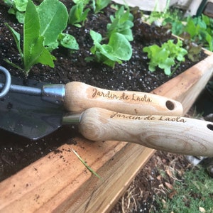 May include: Two wooden-handled gardening tools with the text "Jardin de Laola" engraved on the handles. The tools are lying on top of a wooden planter box filled with soil and green leafy plants.