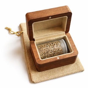 Può includere: Una piccola scatola di legno aperta con interno color chiaro e un contenitore di plastica trasparente pieno di glitter dorati. La scatola è appoggiata su un sacchetto di iuta beige con un cordoncino dorato.