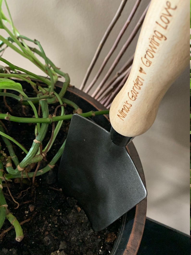 May include: A black gardening trowel with a wooden handle. The handle is engraved with the text "Nina's Garden &hearts; Growing Love".