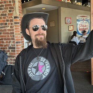 May include: A person wearing a black jacket, a black t-shirt with a pink star and the words "Joyful Deviancy" in a circle, and a brown cowboy hat. They are taking a selfie with a black smartphone.