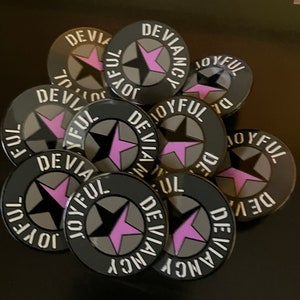 Può includere: Primo piano di diversi spilli circolari neri e grigi con una stella rosa e le parole "Joyful Deviancy" in lettere bianche.