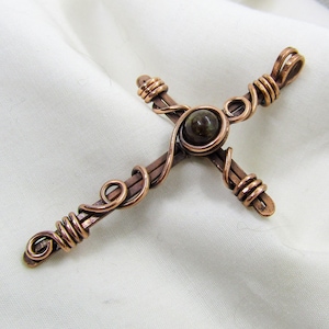 May include: A handcrafted copper cross pendant with a dark green stone at the center. The cross is made of copper wire wrapped around the arms and center, with decorative swirls. The pendant is displayed on a white fabric.