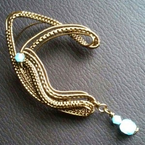 May include: A gold wire ear cuff with two light blue gemstones. The cuff has a curved shape and a dangling gemstone.