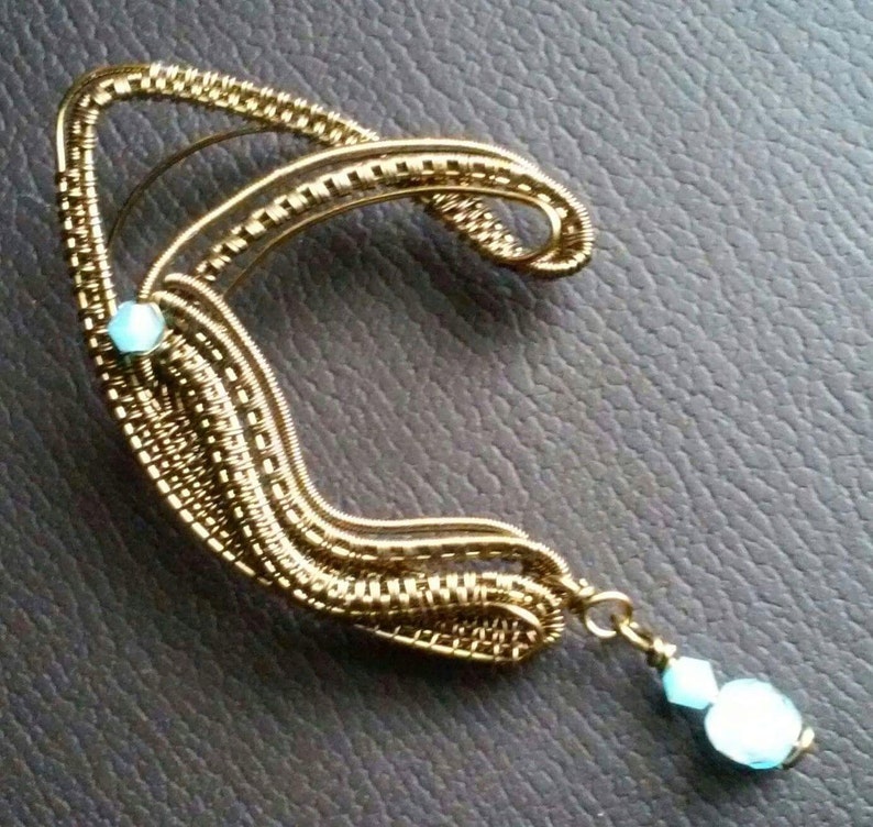 May include: A gold wire ear cuff with two light blue gemstones. The cuff has a curved shape and a dangling gemstone.