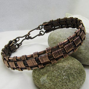 May include: A handcrafted copper wire bracelet with a woven design. The bracelet features a series of interwoven copper wires, creating a textured, openwork pattern. The bracelet is displayed on two gray stones, showcasing its intricate craftsmanship.