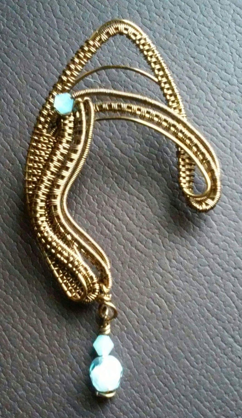 May include: A gold wire wrapped ear cuff with two light blue crystal beads. The ear cuff is shaped like a crescent moon and has a delicate, intricate design.